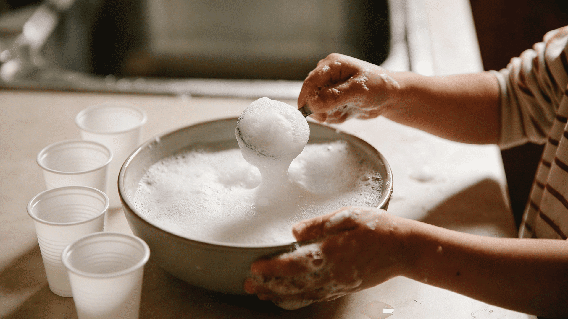 a child uses a metal scoop to lift a large mound of white soap suds from a bowl next to small plastic cups.