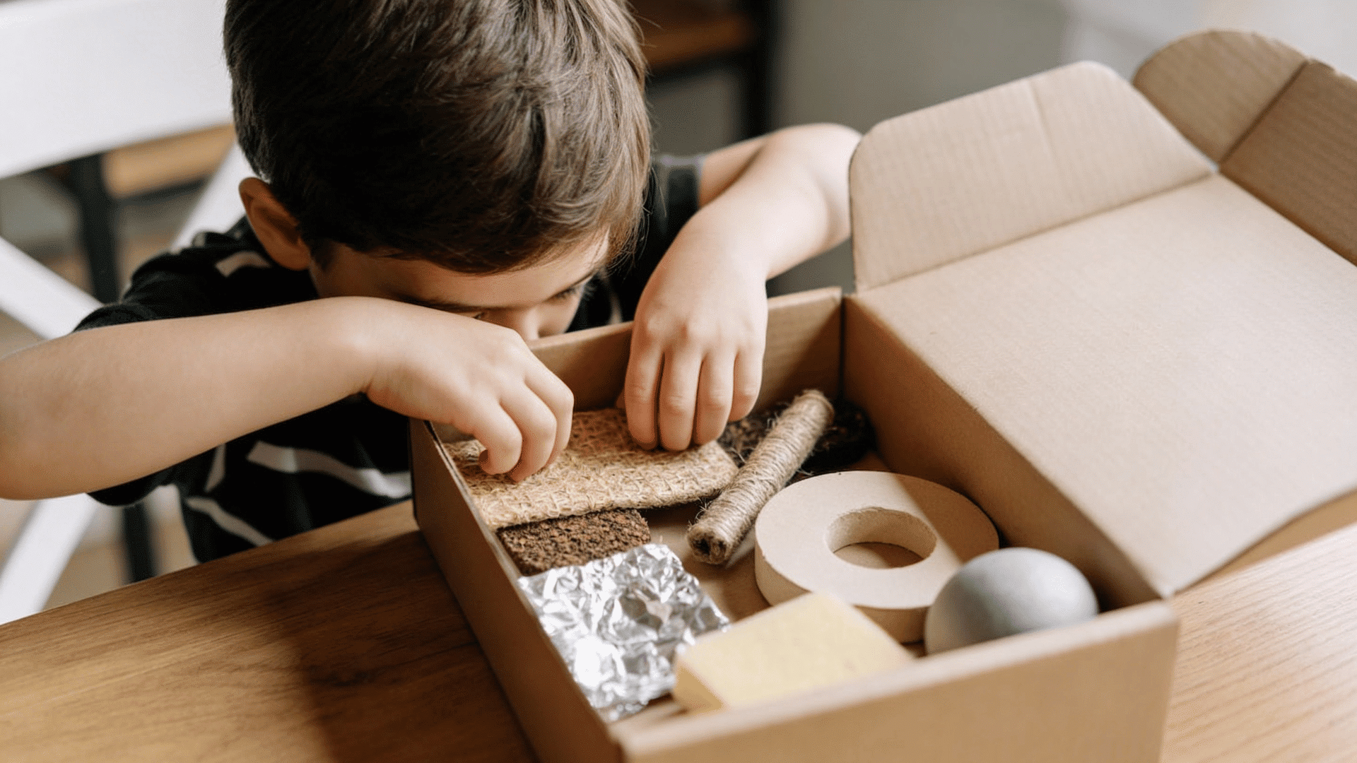 a curious boy leans over a cardboard box, feeling different textures like rope, sponge, and crinkled foil.