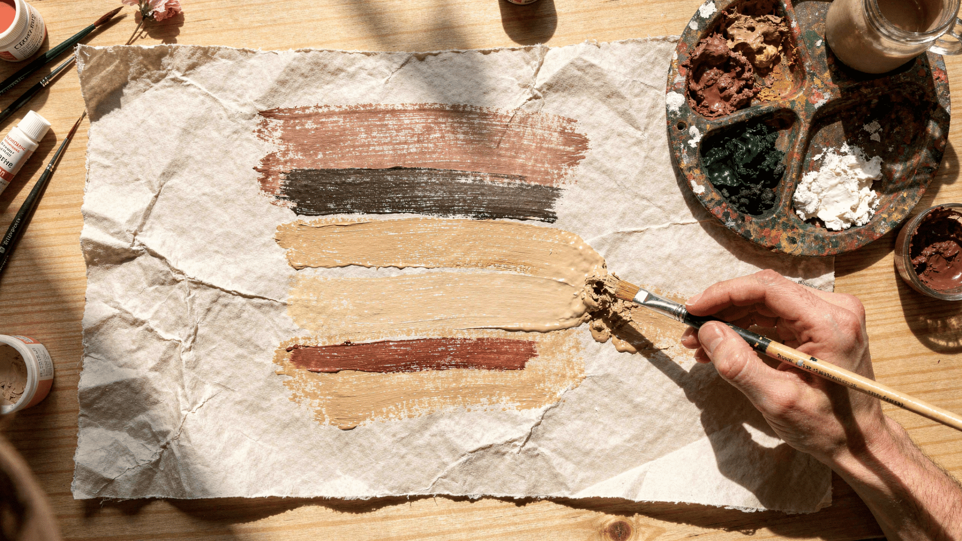 a hand uses a brush to create thick, earth-toned gouache swatches on a piece of wrinkled, primed paper.