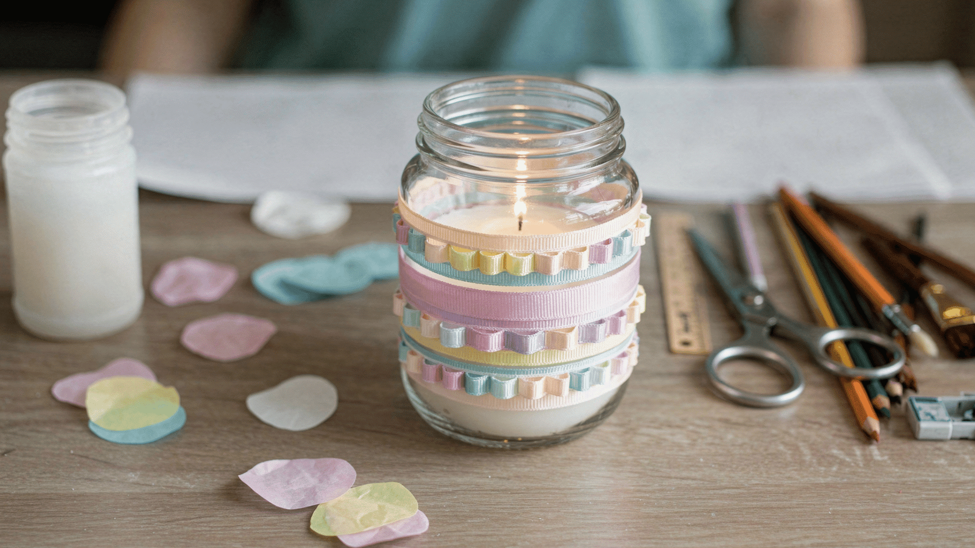 a lit candle sits inside a glass jar decorated with colorful ruffled ribbons on a wooden desk with craft supplies.