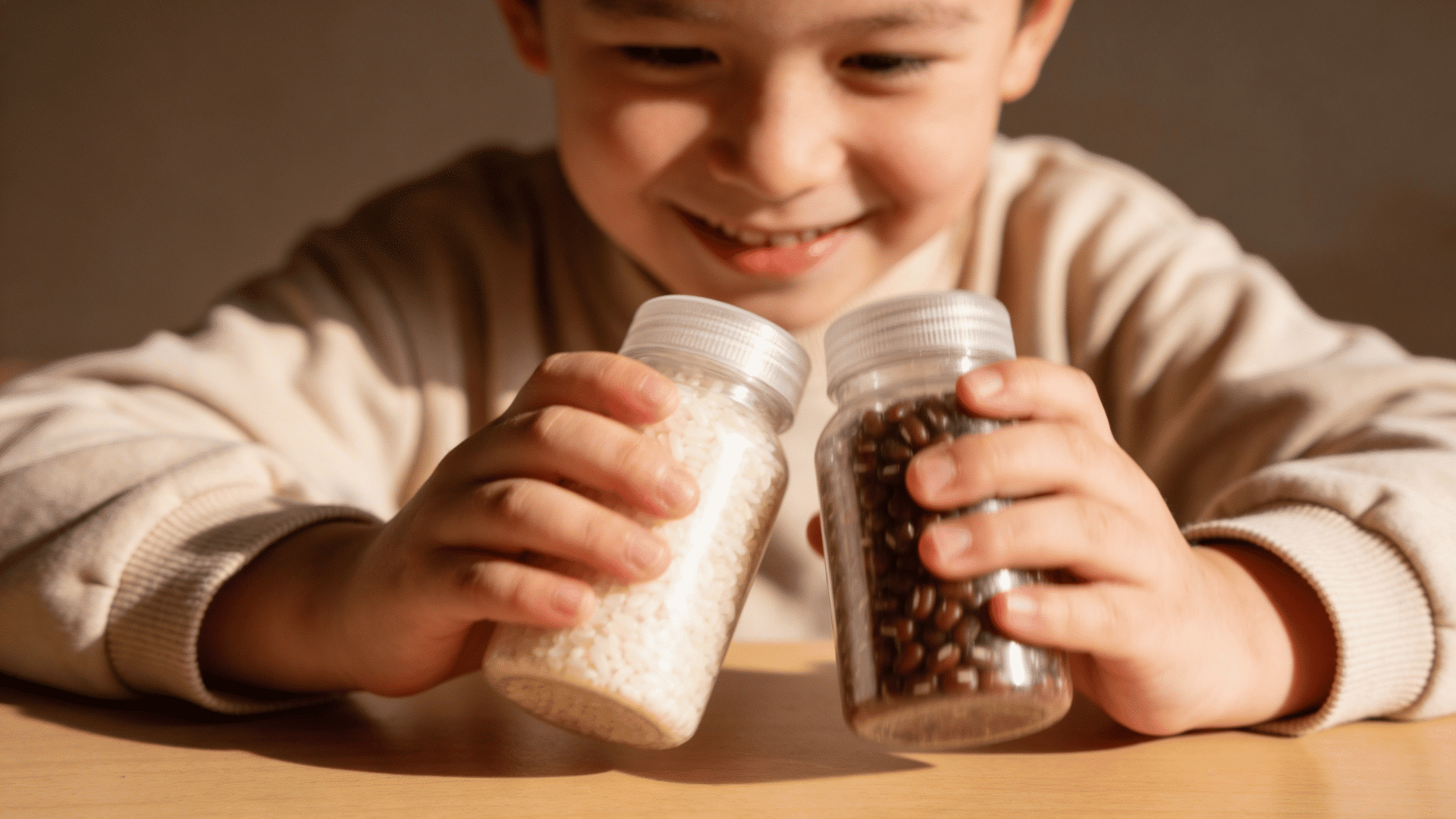 a smiling boy holds two clear plastic bottles filled with white rice and red beans to compare their sounds. (1)