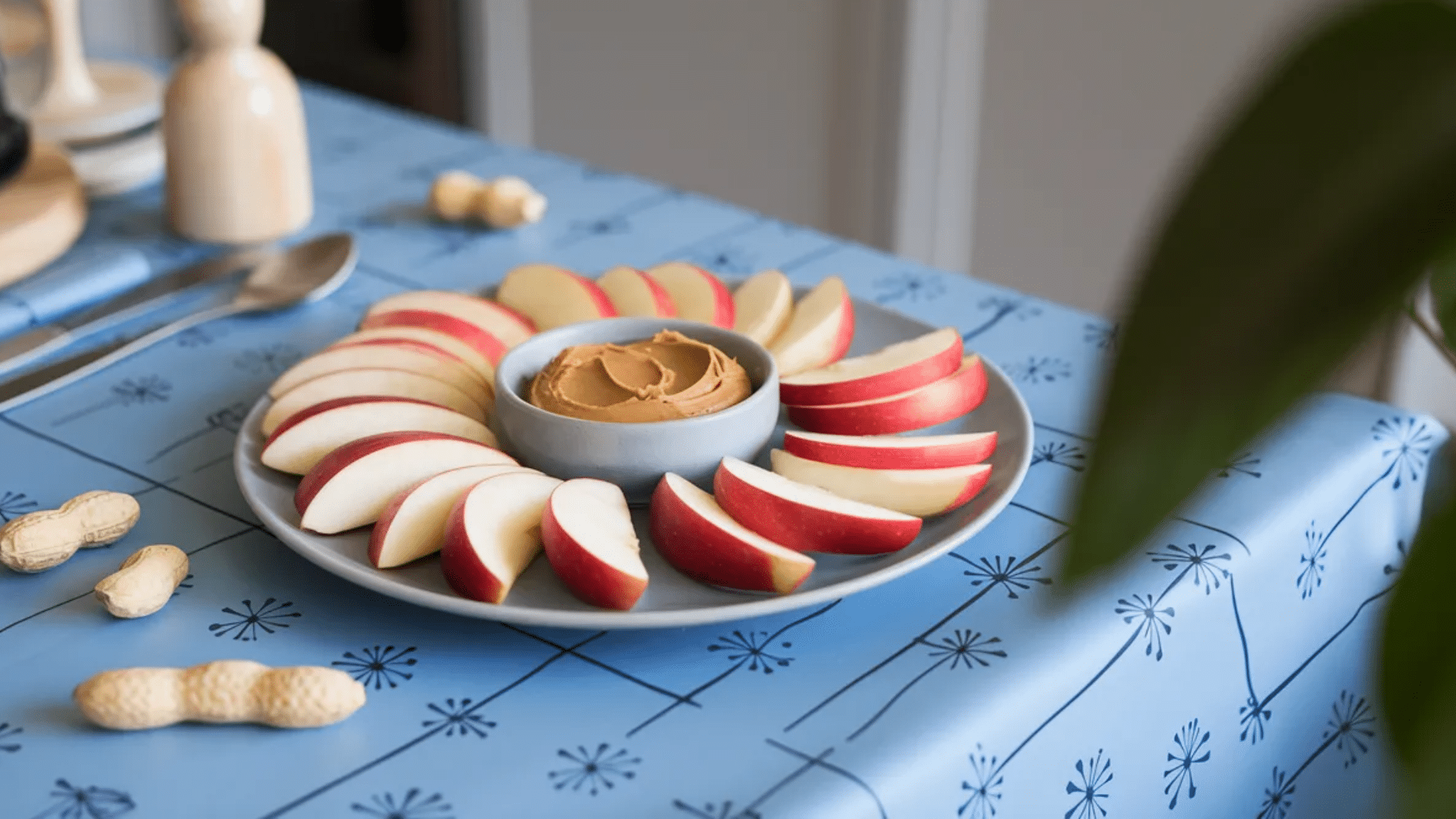 apple slices arranged in circle around small bowl of peanut butter on plate with whole peanuts and spoon on blue tablecloth