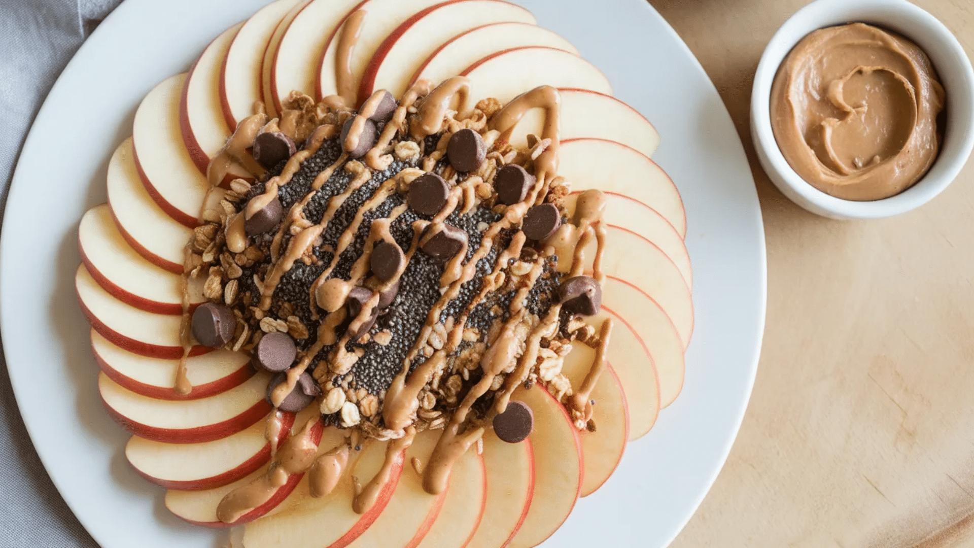 apple slices arranged in circle topped with granola chia seeds chocolate chips and peanut butter drizzle on white plate with peanut butter dip nearby