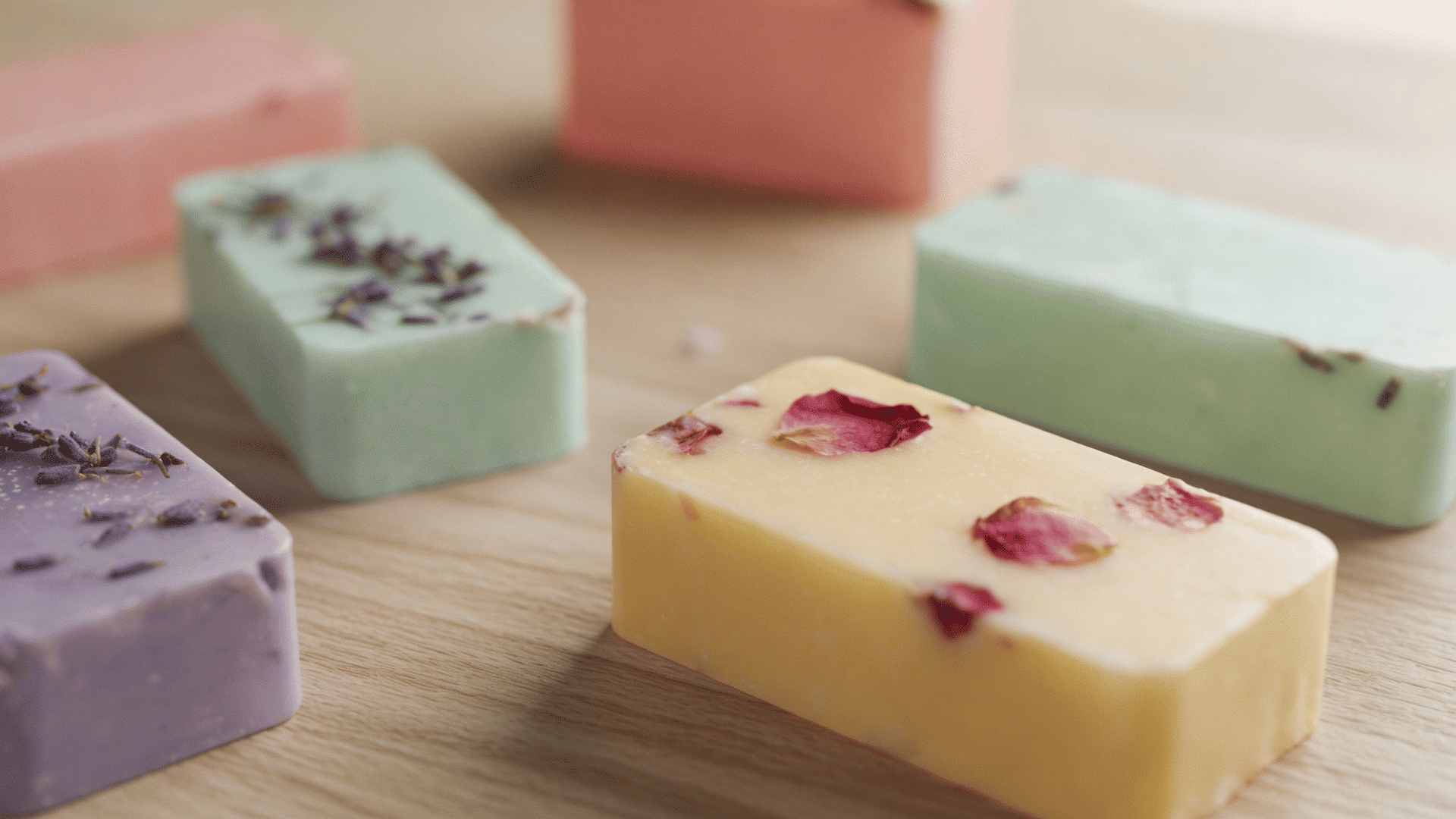 assorted handmade rectangular soap bars in pastel colors decorated with dried lavender and rose petals on a wooden surface.