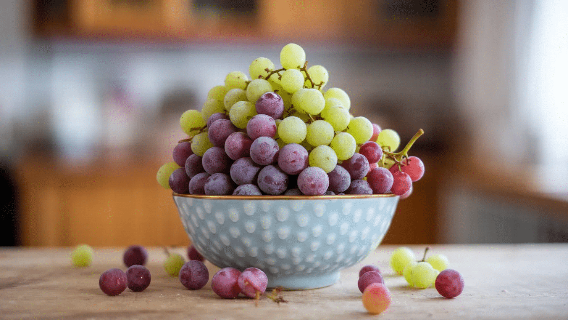 bowl filled with green and red grapes piled high on kitchen table with a few loose grapes scattered around the bowl
