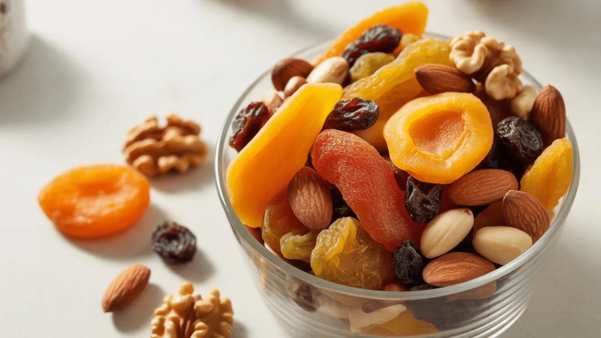bowl of trail mix with dried apricots raisins almonds walnuts and peanuts with a few scattered nuts and dried fruit on table