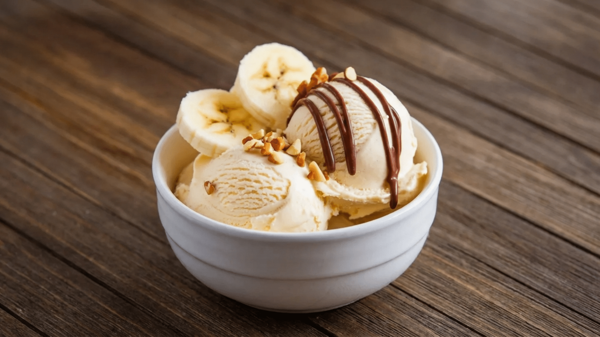 bowl of vanilla ice cream topped with banana slices chopped nuts and chocolate drizzle placed on rustic wooden table