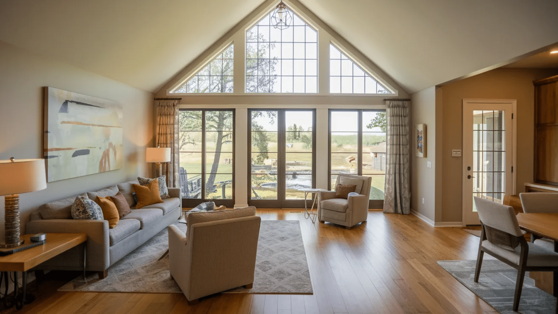 bright living room with vaulted ceiling and large triangular windows, gray sofa and chairs, wooden floor, and wide glass doors