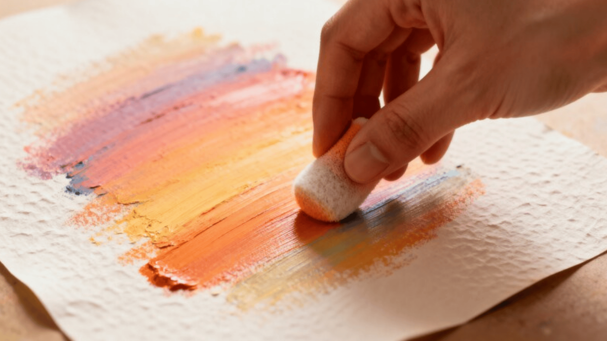 close-up of a hand using a pastel stick to blend soft colors of orange on paper