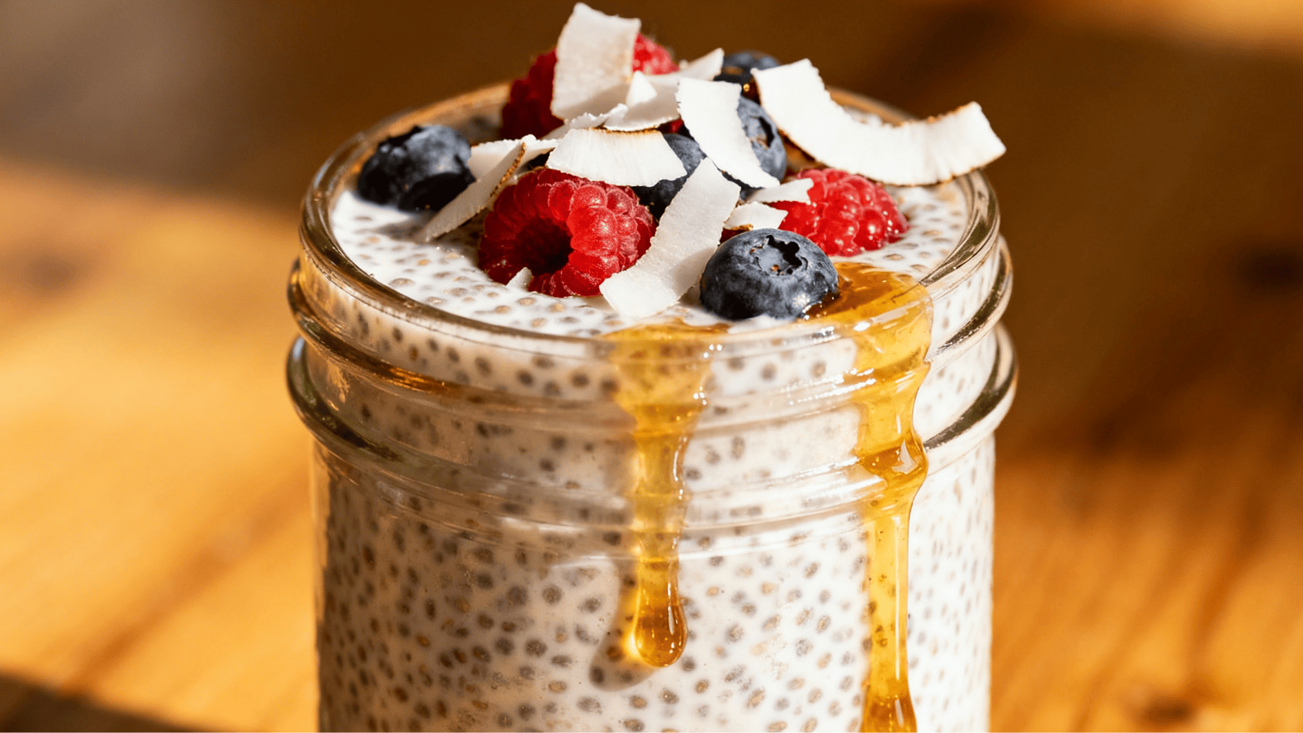 coconut chia pudding in a glass jar topped with blueberries raspberries coconut flakes and honey dripping down the side on wooden table