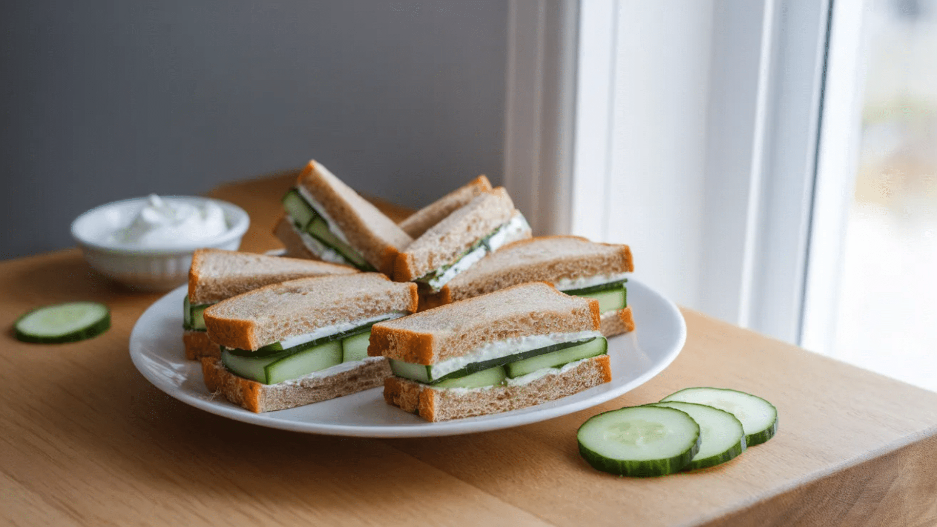 cucumber tea sandwiches with cream cheese on whole wheat bread arranged on white plate with cucumber slices on wooden table