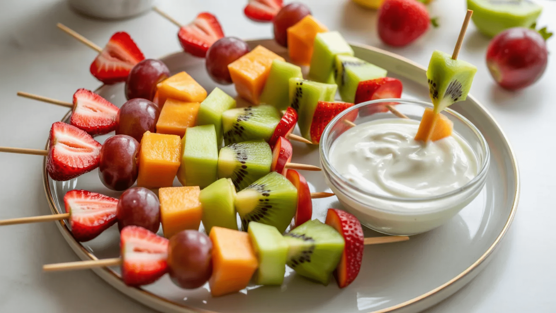 fruit skewers with strawberries grapes kiwi melon and cantaloupe arranged on plate beside bowl of creamy yogurt dip
