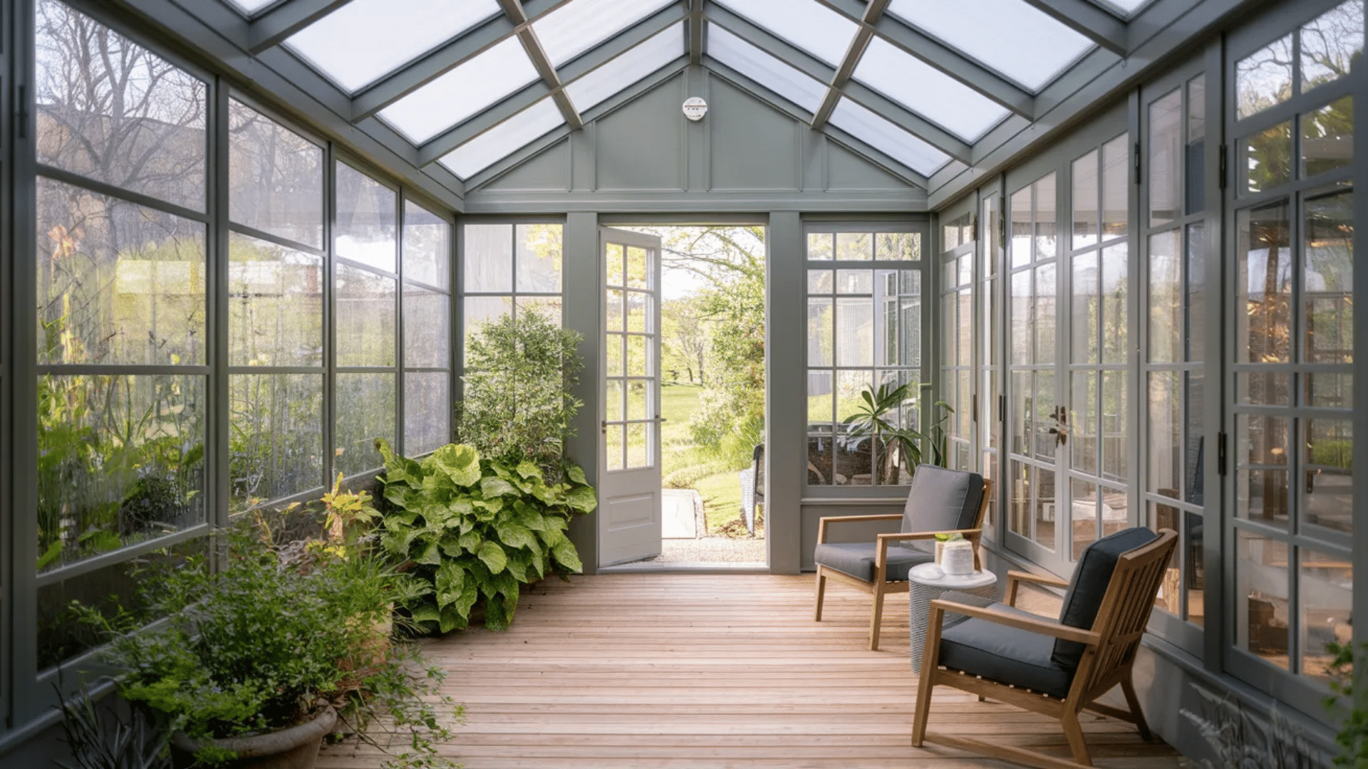 glass sunroom with skylight roof wooden floor lounge chairs indoor plants and open door to garden outside view