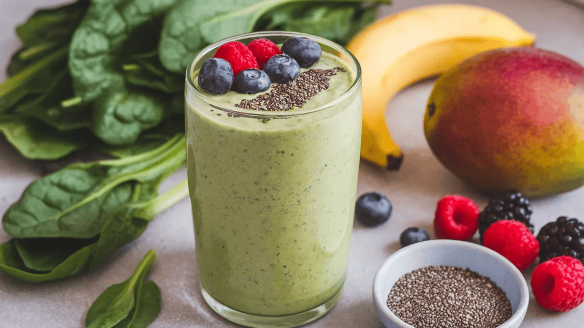 green smoothie topped with chia seeds, raspberries, blueberries, and surrounded by fresh spinach, banana, and mango