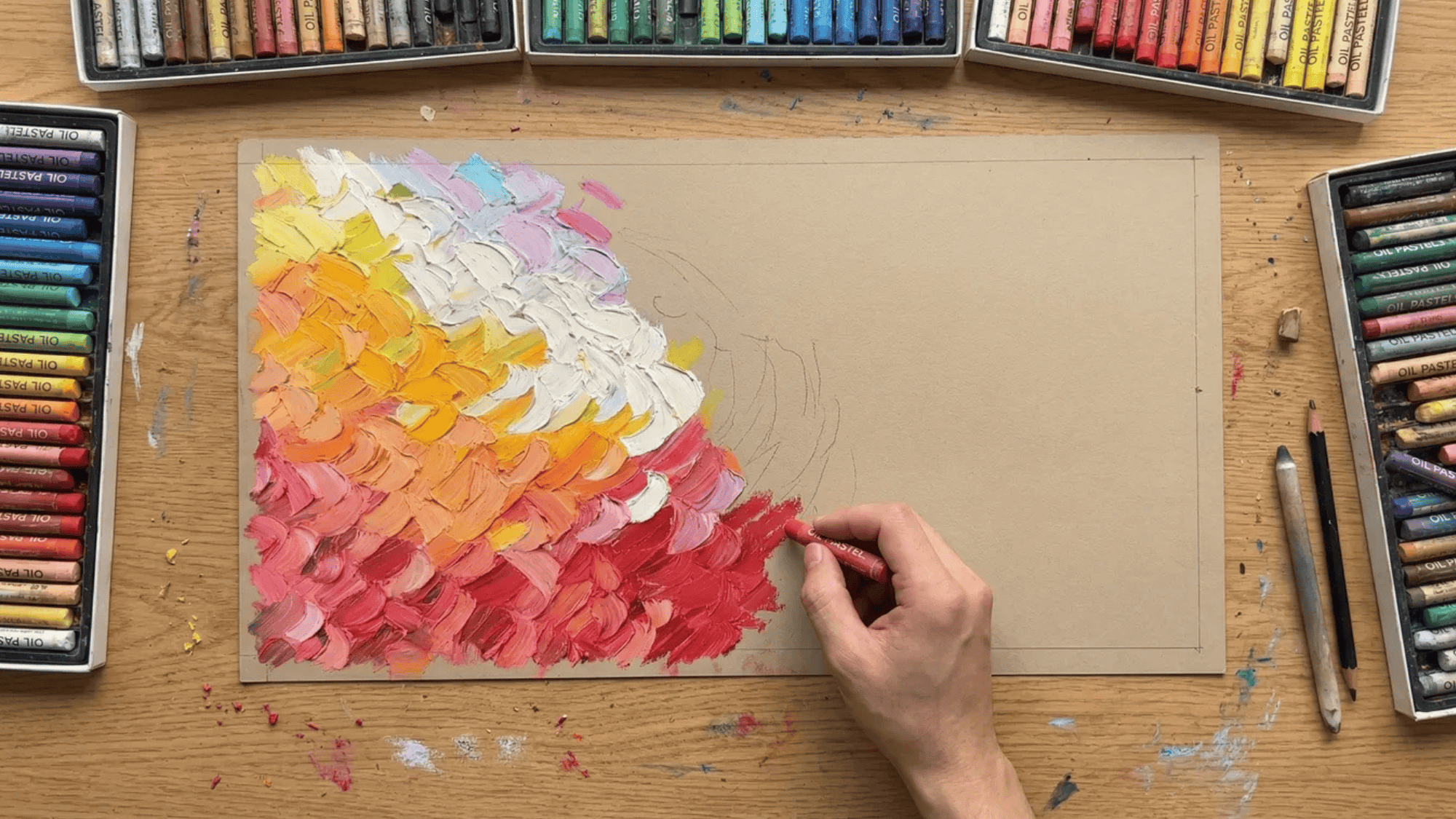 hand using an oil pastel to add red to a colorful pastel artwork, with vibrant yellow, pink, and red hues on textured paper, surrounded by oil pastel boxes