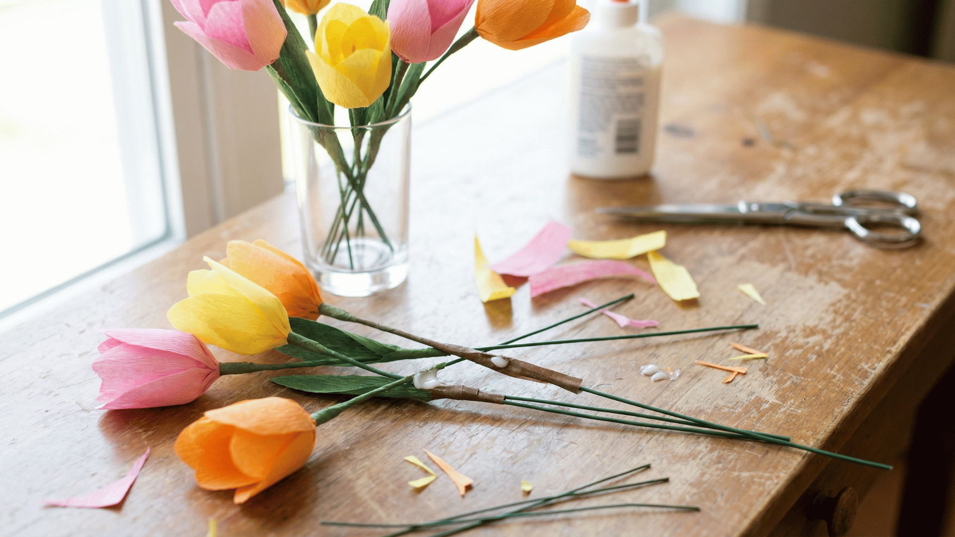 handmade crepe paper tulips in pink, yellow, and orange lie on a wooden table next to scissors and a vase of flowers.