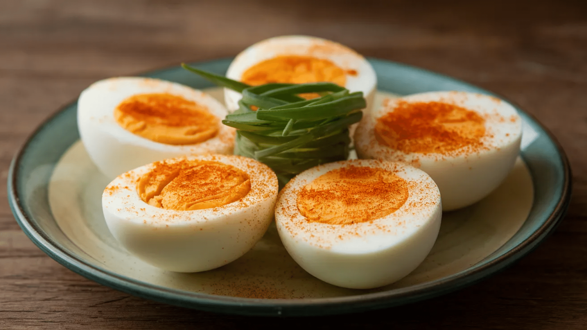 hard boiled egg halves sprinkled with paprika arranged on plate with small bundle of green chives in center on wooden table
