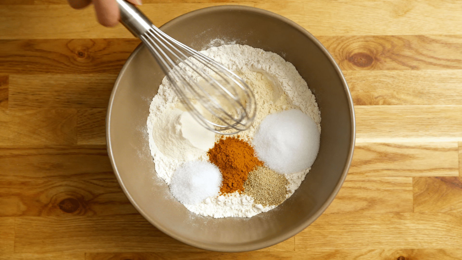 overhead view of flour, baking soda, baking powder, pumpkin spice, and salt being whisked together in a mixing bowl on a wooden counter.