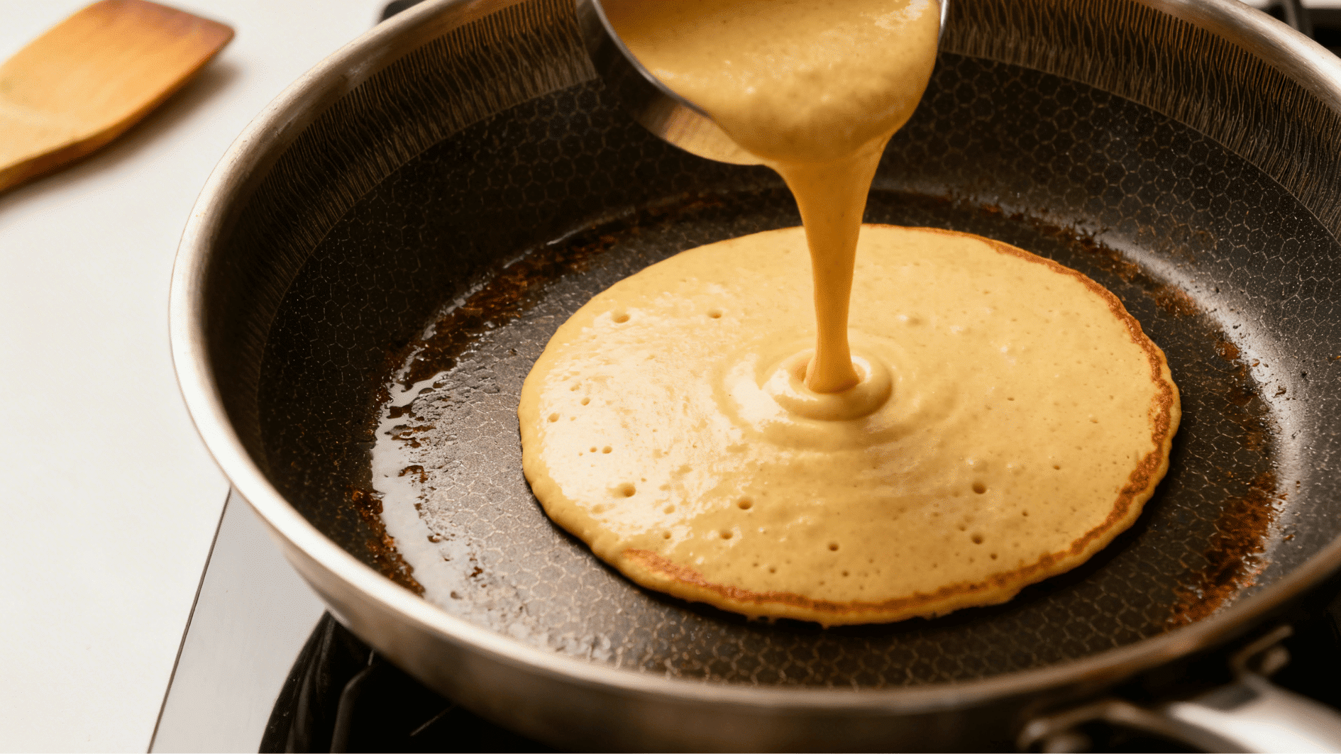 pancake batter being poured onto a hot nonstick pan forming a round shape as it begins cooking for homemade pancakes