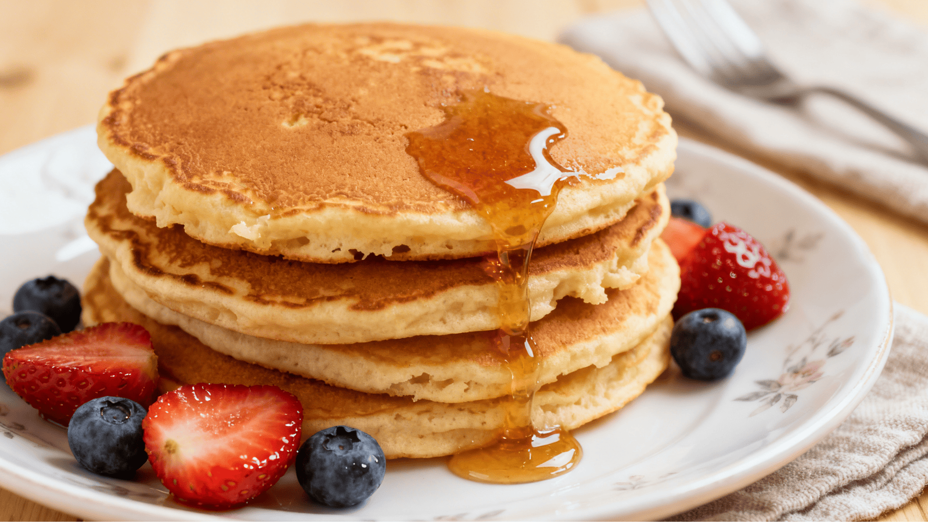 pancakes stacked on a plate with syrup dripping over the sides and fresh strawberries and blueberries beside the pancakes
