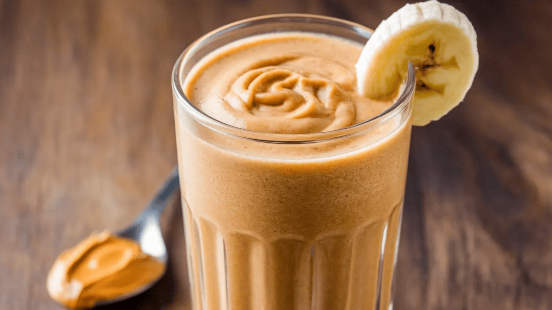 peanut butter banana smoothie in a glass, garnished with a banana slice, with a spoon of peanut butter beside it