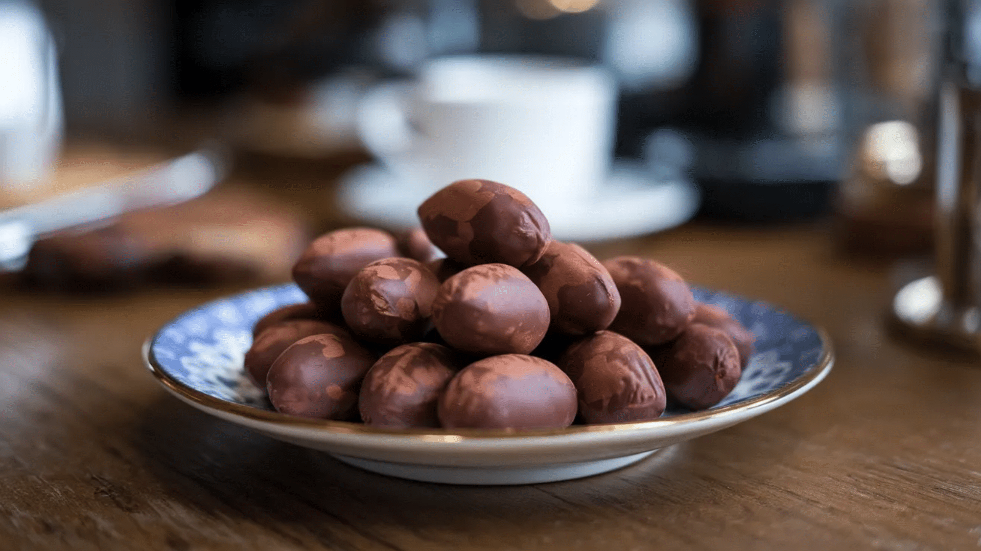 plate of chocolate covered nuts stacked in small pile on wooden table with blurred coffee cup and cafe setting in background