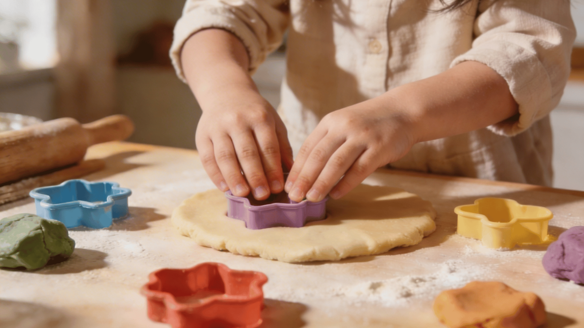 playdough cutters a child’s hands use a purple flower-shaped cutter on beige playdough next to colorful dough and a rolling pin.