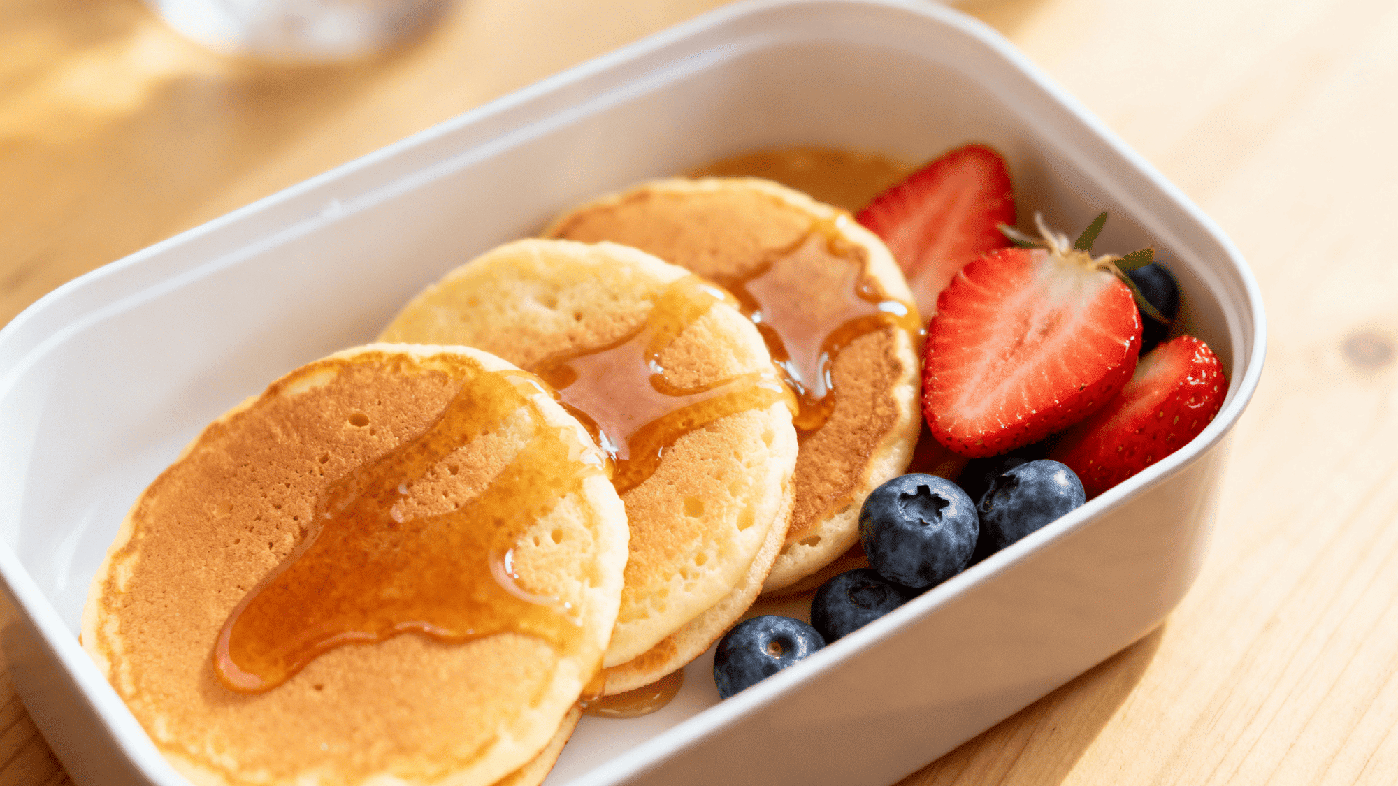 small pancakes arranged in a lunchbox with sliced strawberries and blueberries, light maple glaze drizzle