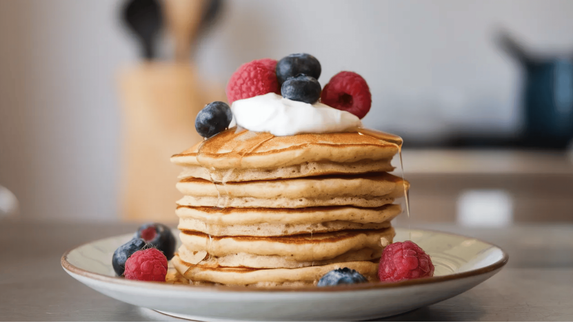stack of pancakes topped with fresh raspberries, blueberries, a dollop of cream, and a drizzle of syrup