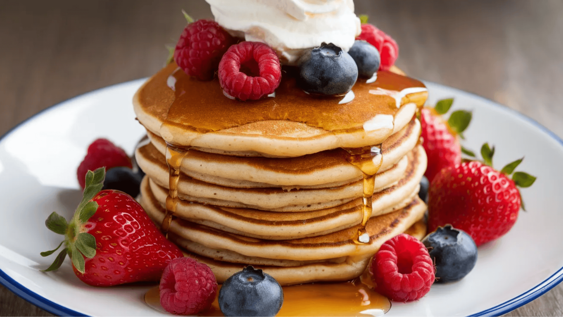 stack of pancakes topped with whipped cream, fresh raspberries, blueberries, strawberries, and maple syrup