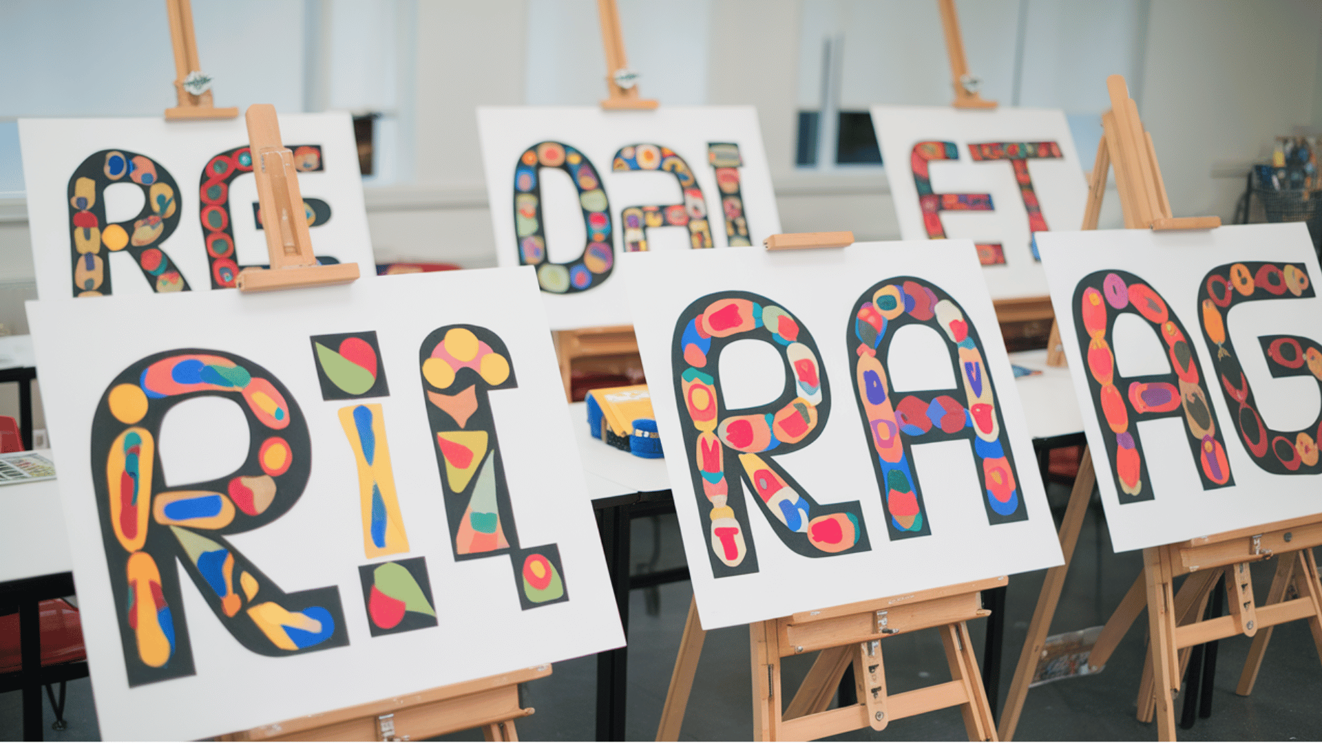 students' colorful abstract name design artwork displayed on easels in an art classroom.