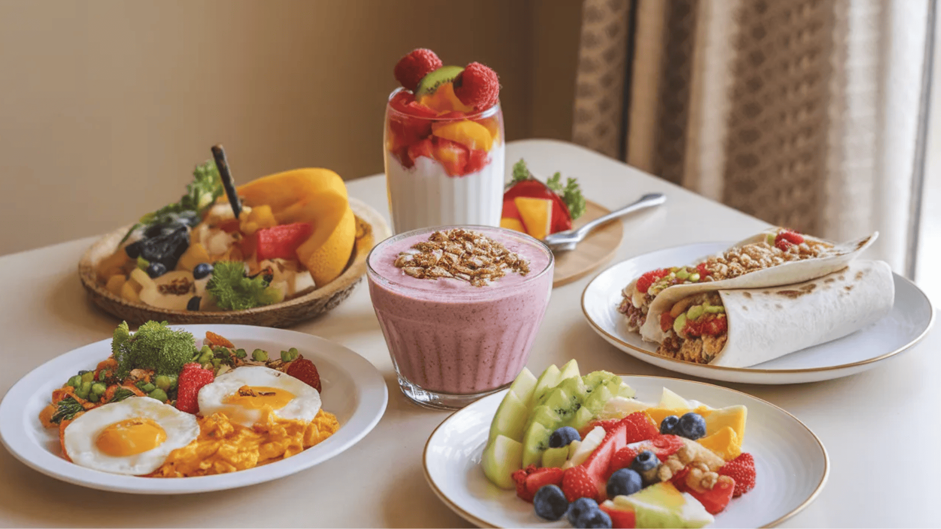 variety of healthy breakfast items including scrambled eggs, fruit salad, smoothies, and a wrap with granola