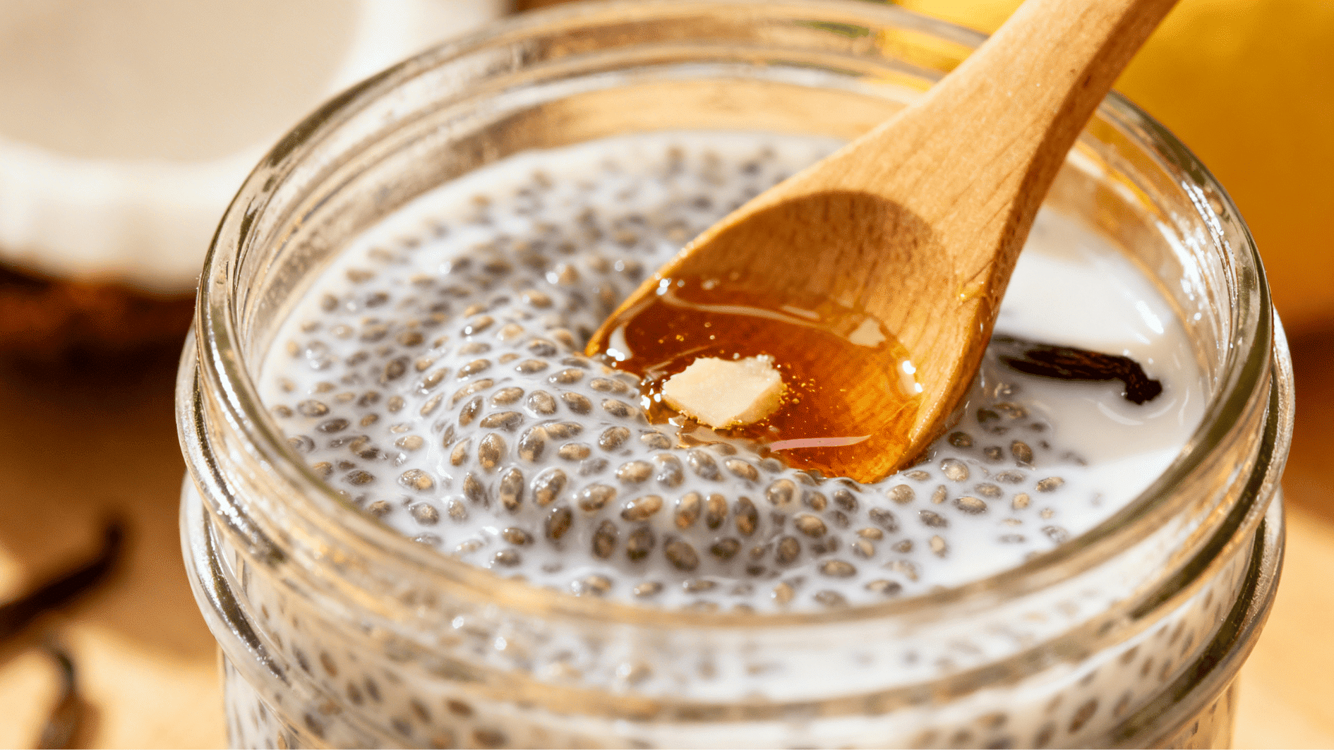 wooden spoon adding maple syrup into a jar of coconut chia pudding mixture with visible chia seeds and vanilla pod in the background