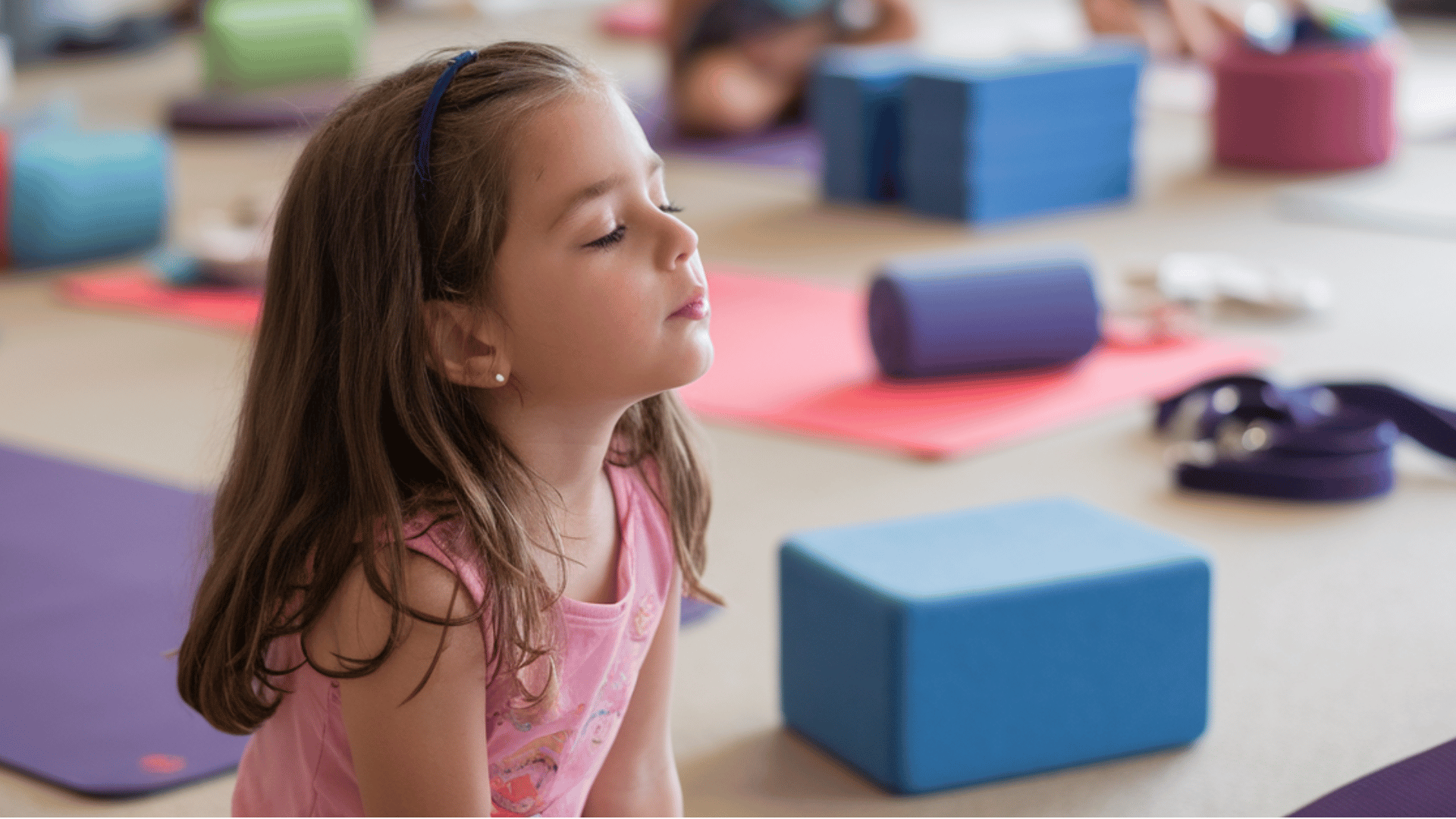a child participates in a yoga class for kids, sitting in a calming position with eyes closed and relaxed.