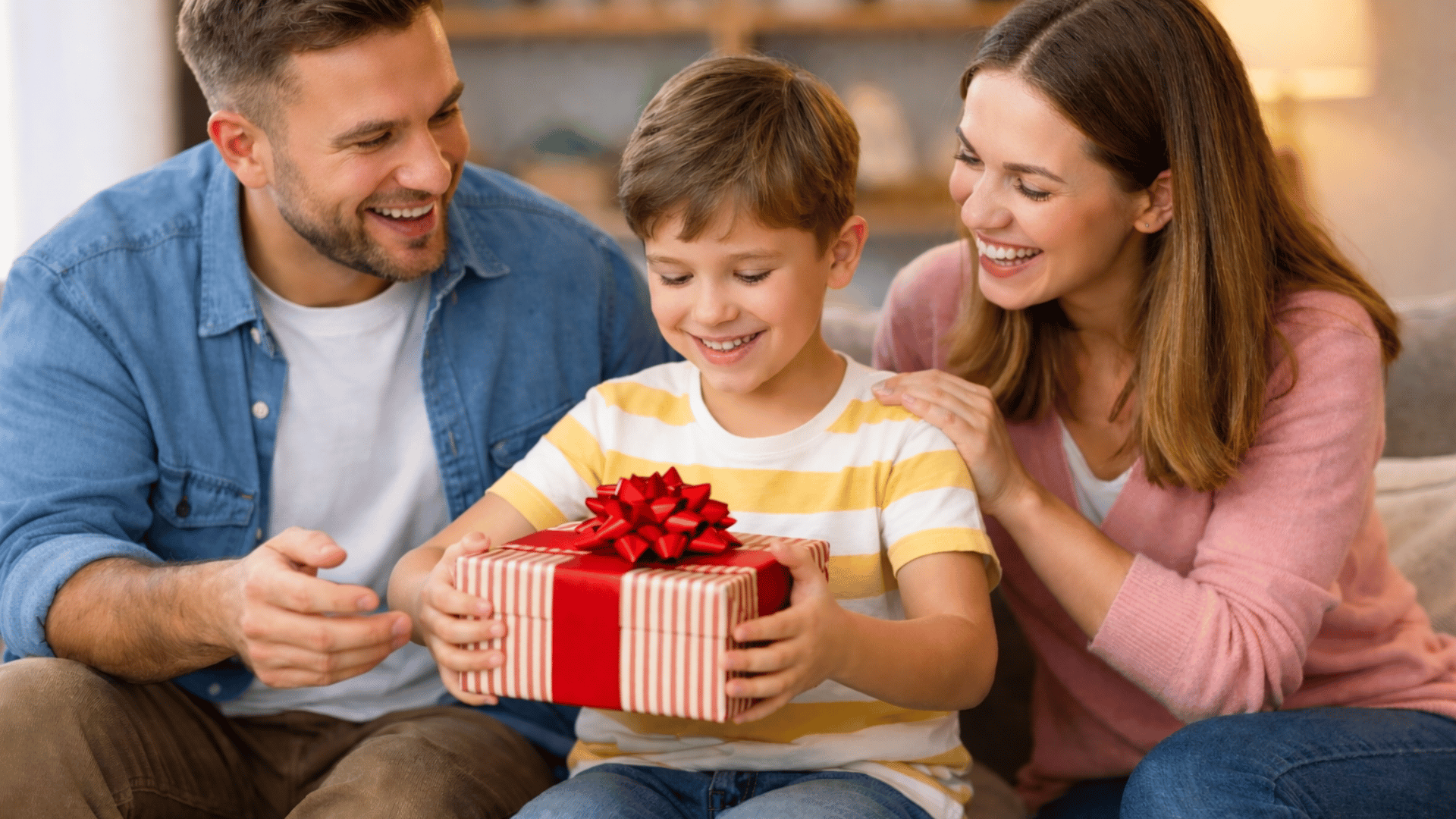 a happy family of three, with parents watching as their son opens a gift on the couch