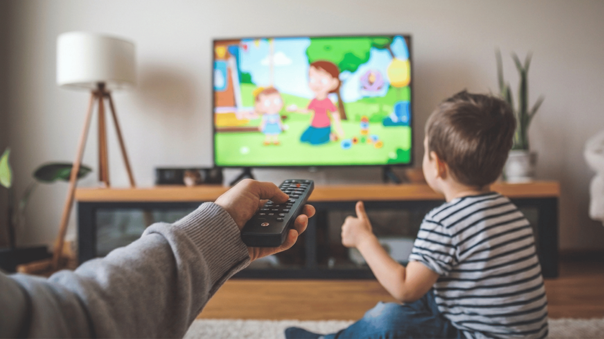 a parent holding a remote to pause a cartoon on tv while a child watches from the floor.