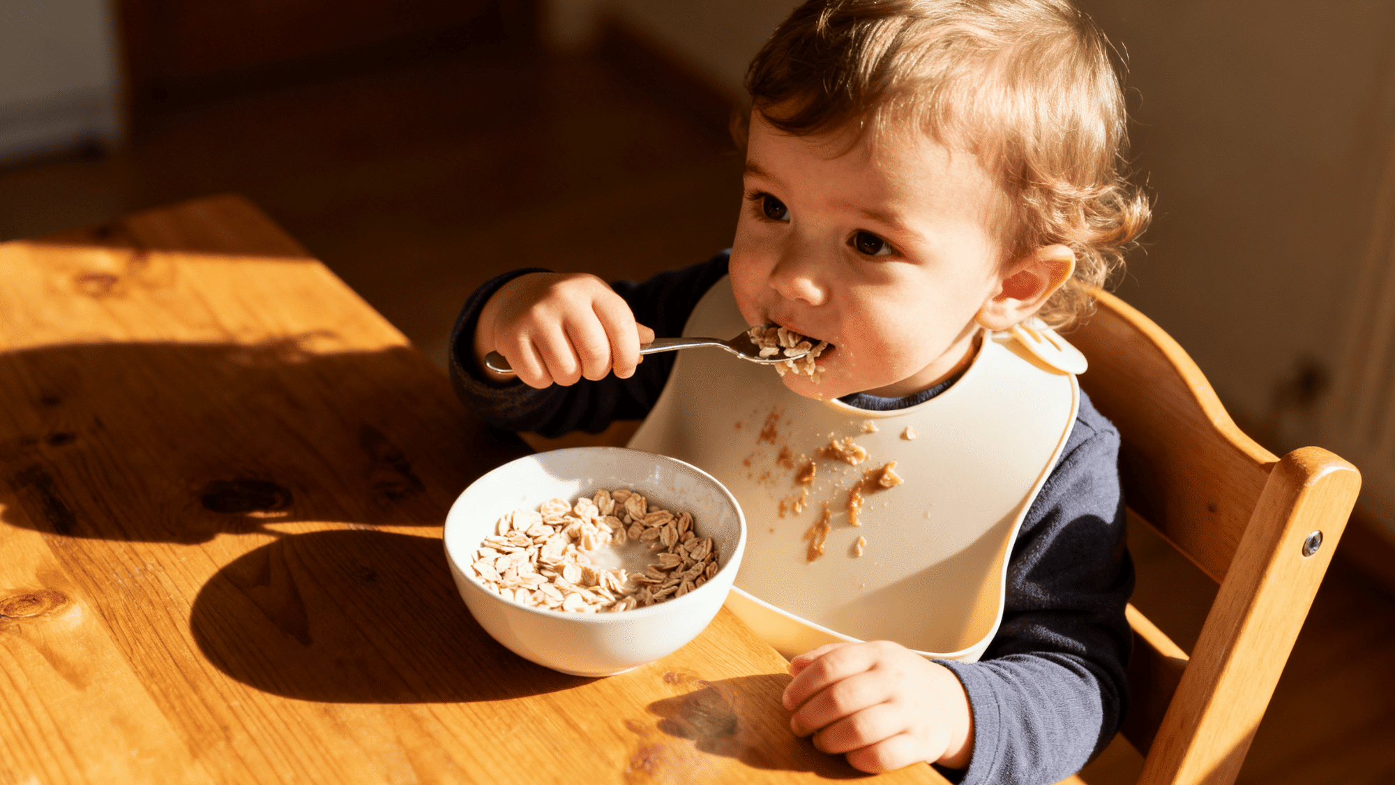 a toddler eating oats at a small wooden table, spoon in hand, messy bib, warm morning light