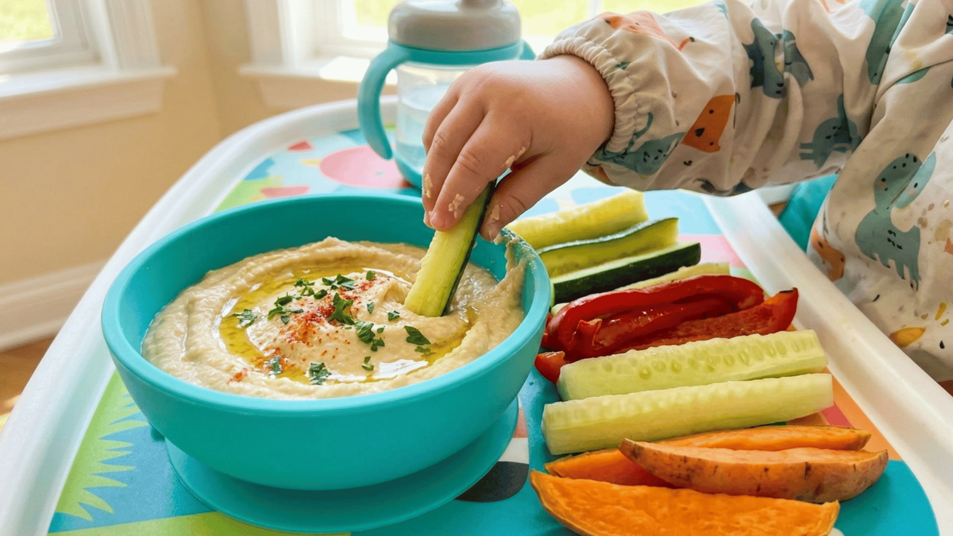 baby dipping cucumber stick into hummus with veggie slices on a colorful tray.