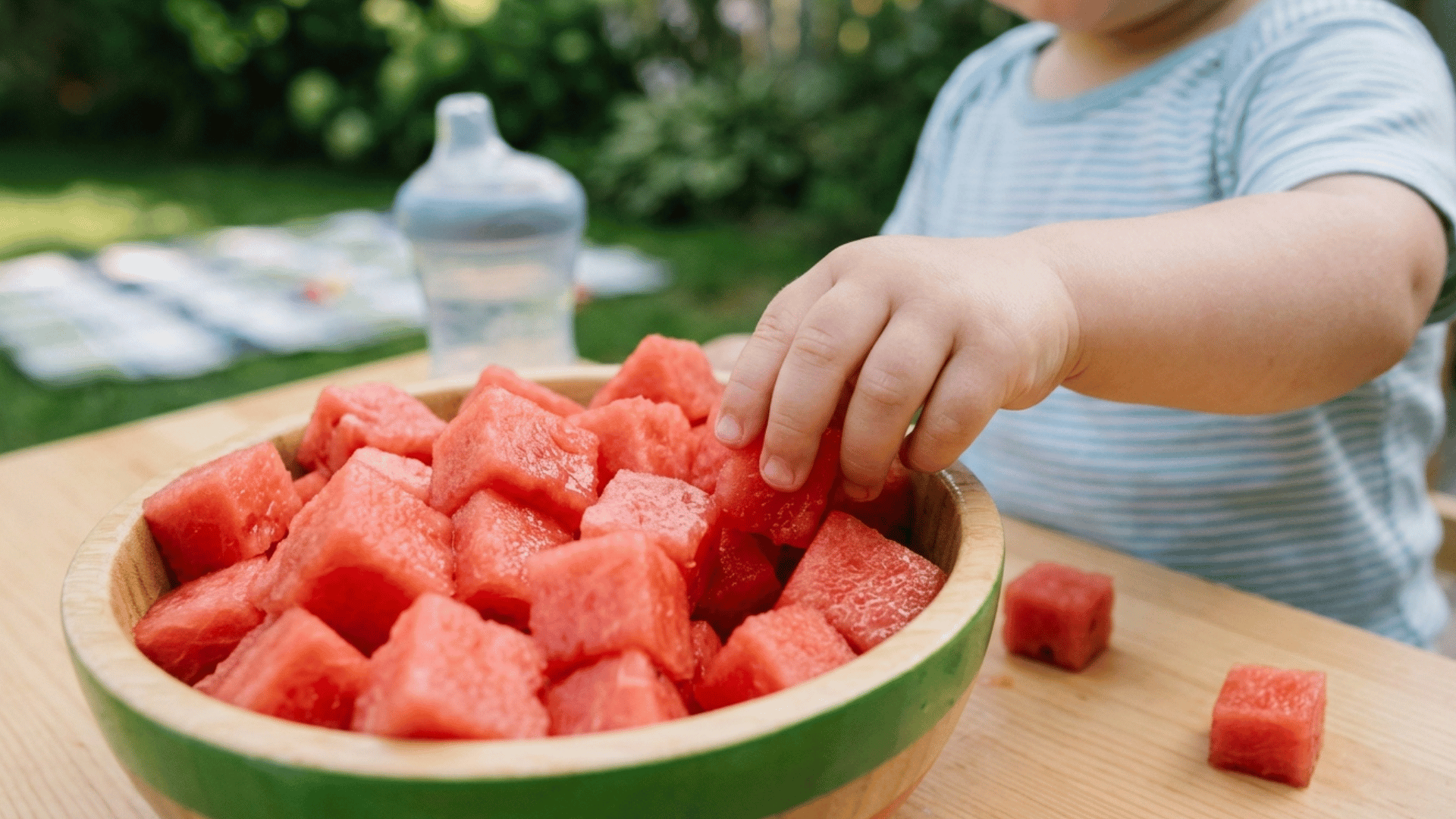 baby hand reaching for fresh watermelon cubes in a bowl outdoors on a sunny day.