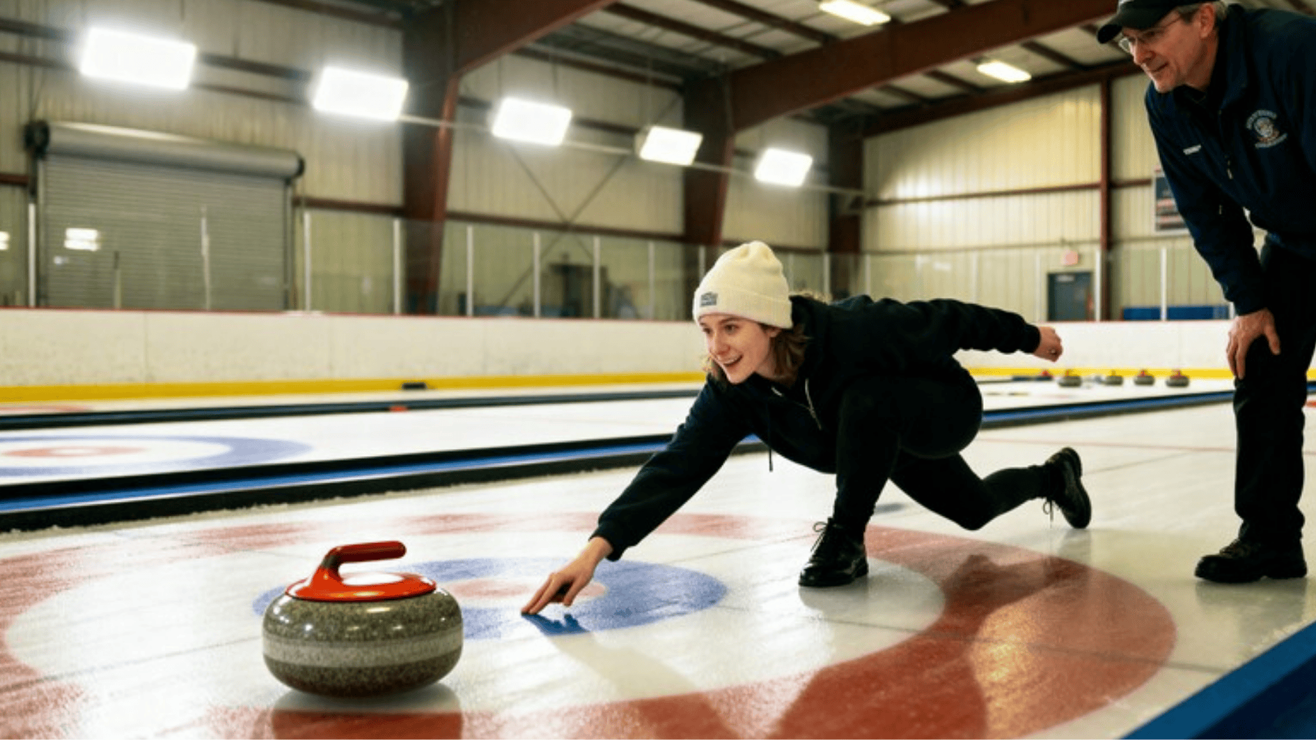 beginner learning to curl on an indoor ice sheet, sliding a stone toward the target, coach watching nearby