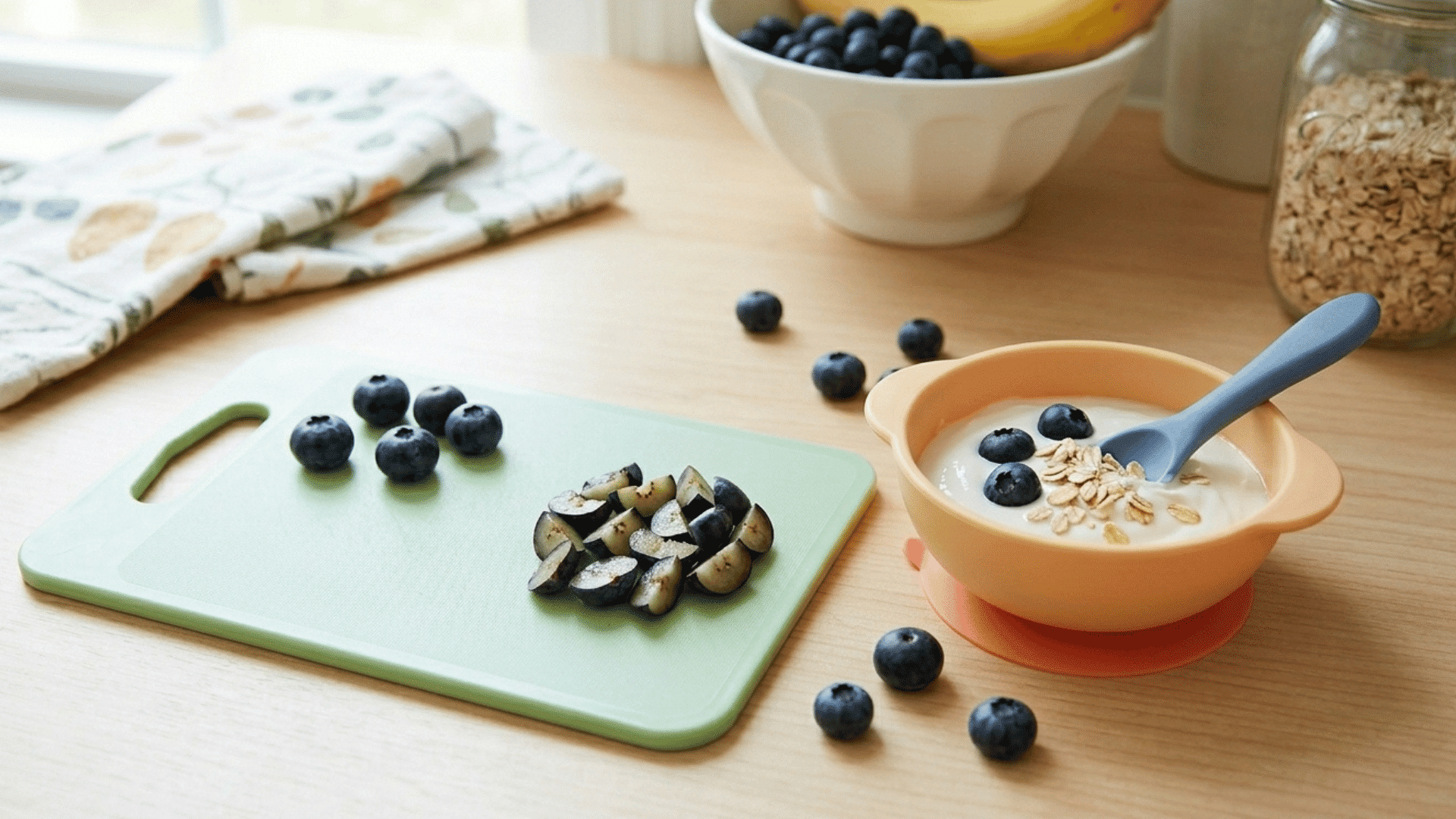 bowl of yogurt with oats and blueberries beside chopped fruit on a cutting board.