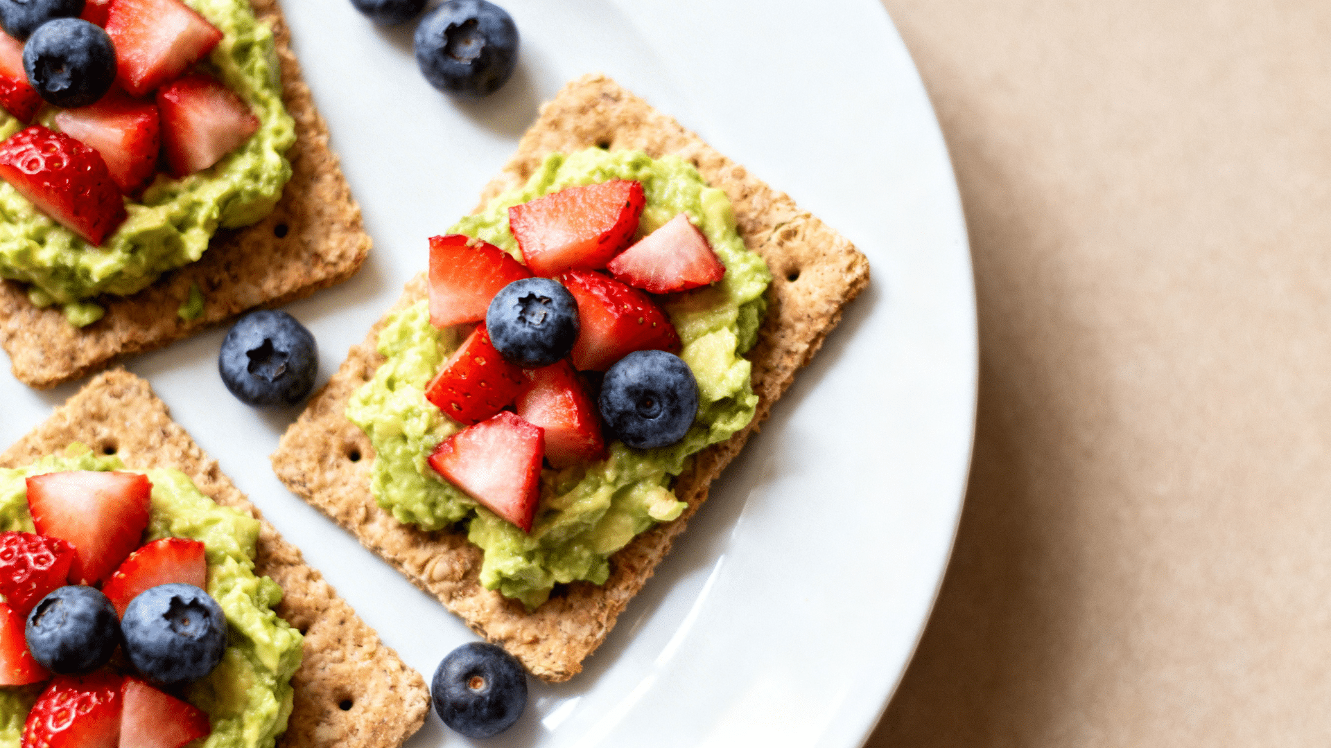 crackers topped with mashed avocado, fresh strawberry slices and blueberries, arranged on a white plate with scattered berries around