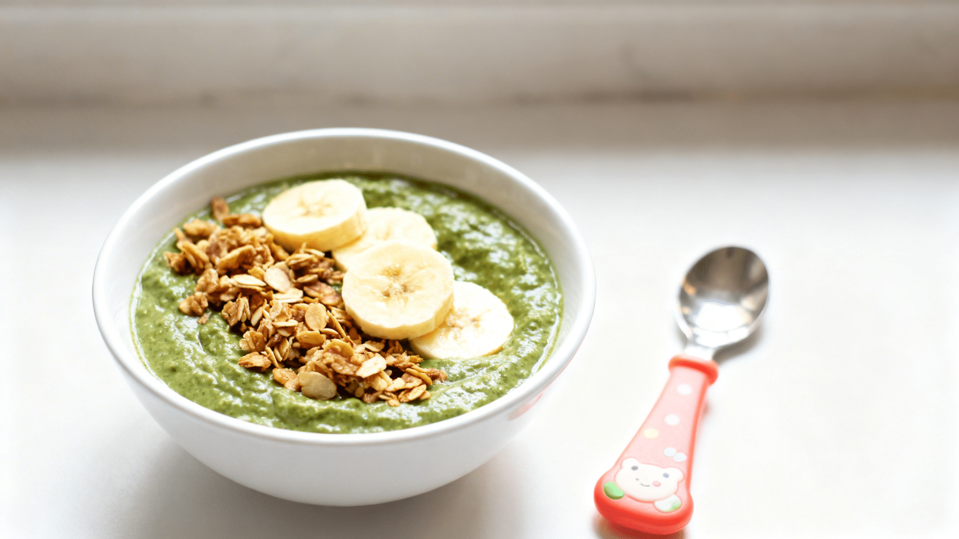 green smoothie bowl topped with banana slices and crunchy granola, placed on a white surface with a small spoon beside it
