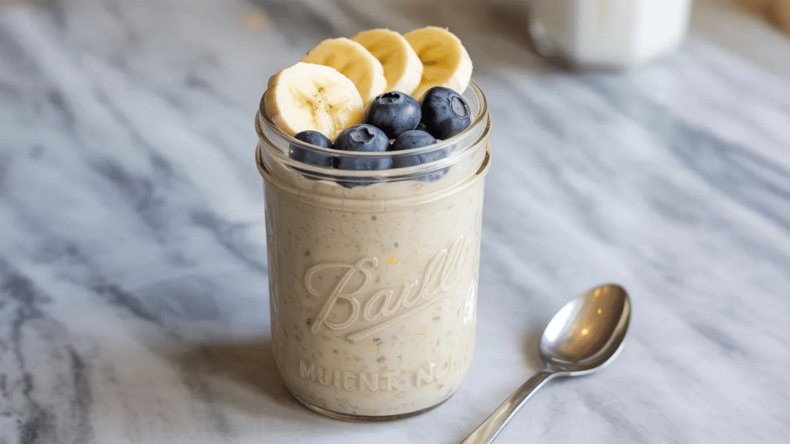 mason jar filled with creamy overnight oats, topped with sliced banana, fresh blueberries, and a drizzle of honey, with a small spoon resting beside it