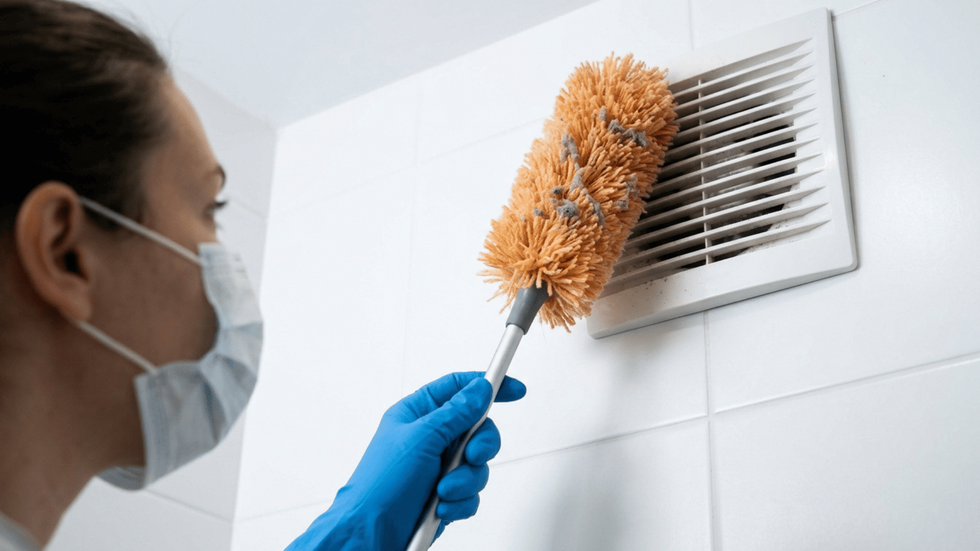 person dusting an air vent with a microfiber duster while wearing gloves and a face mask.