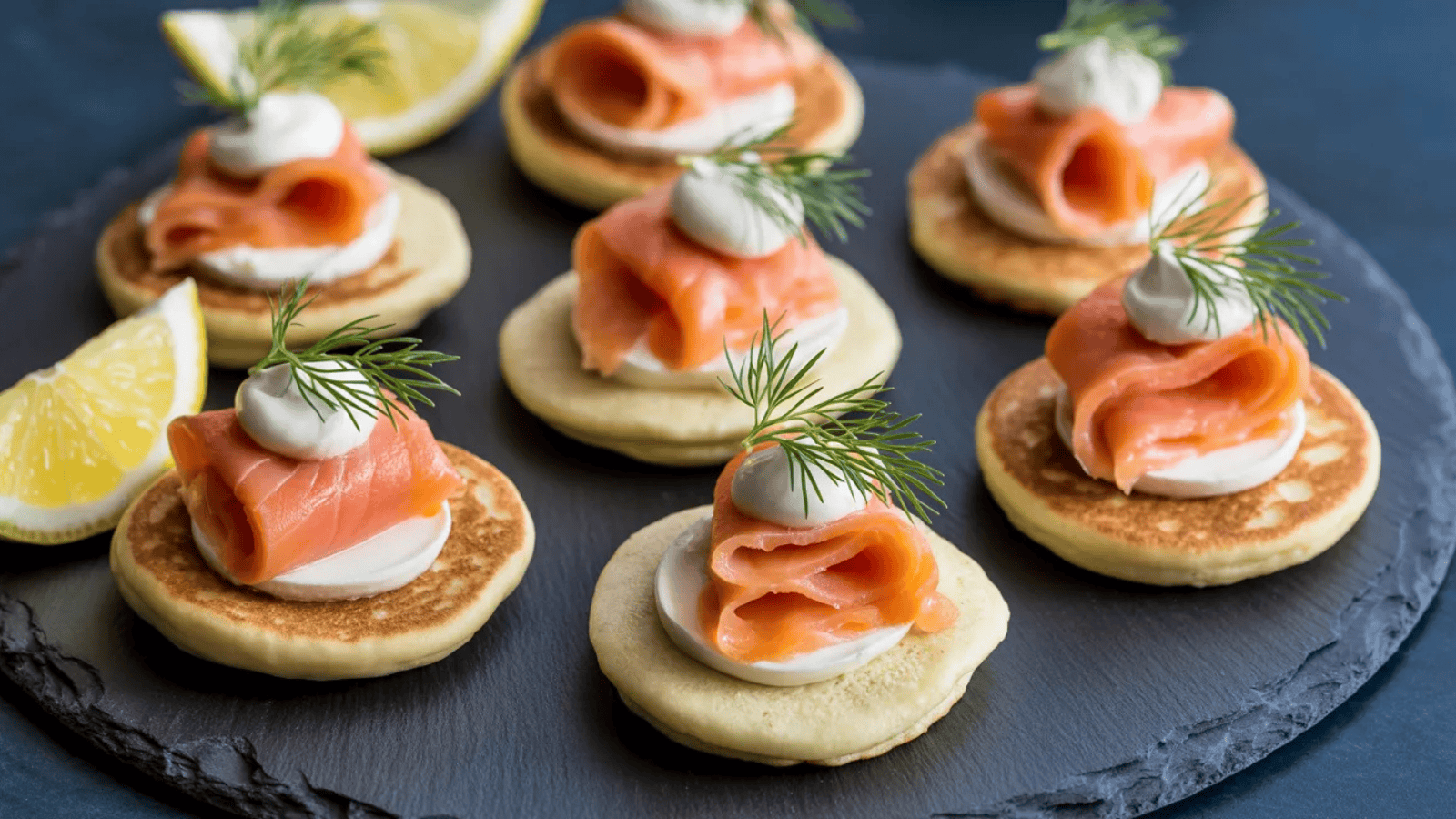 six small golden blini arranged on a slate board, each topped with cream cheese, folded smoked salmon, a tiny spoonful of crème fraîche