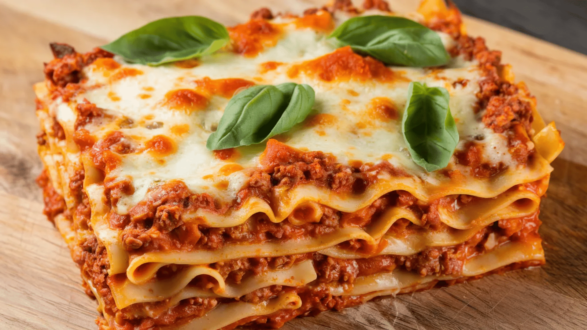 slice of lasagna layered with pasta meat sauce and melted cheese topped with fresh basil on a wooden board
