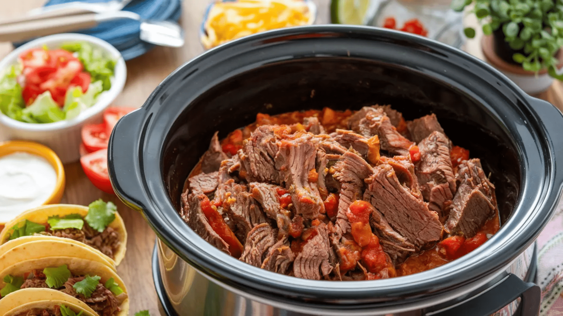 slow cooked shredded beef in a crockpot with tomatoes served with taco ingredients and topping around the dish