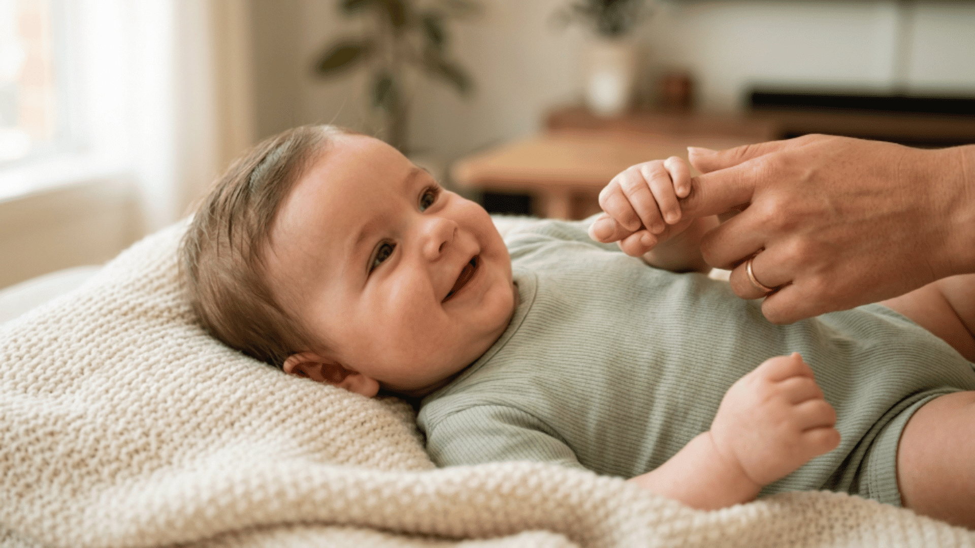 smiling baby lying on a soft blanket while gently holding an adult’s finger with a tiny hand