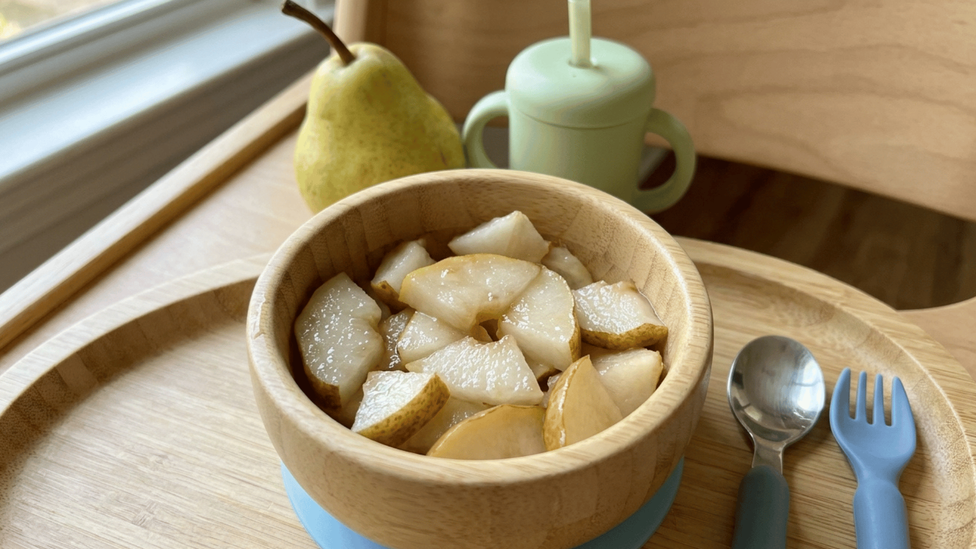 soft pear slices in a wooden bowl with baby cup and utensils on a tray by the window.